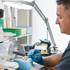 Doświadczony technik dentystyczny w niebieskich rękawiczkach precyzyjnie pracuje nad modelem zębów pod lampą.