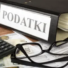 Segregator z napisem "PODATKI", kalkulator, okulary i dokumenty. Obok leży moneta, symbolizująca potencjalną kwotę zmniejszającą podatek.