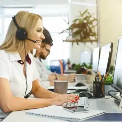 Uśmiechnięta blondynka w słuchawkach z mikrofonem pracuje jako dyspozytor. W tle widać innych pracowników i monitory komputerów.