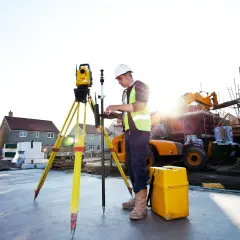 Geodeta w kamizelce odblaskowej pracuje z niwelatorem na budowie.
