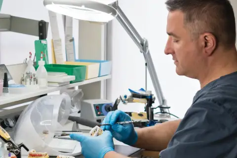 Doświadczony technik dentystyczny w niebieskich rękawiczkach precyzyjnie pracuje nad modelem zębów pod lampą.