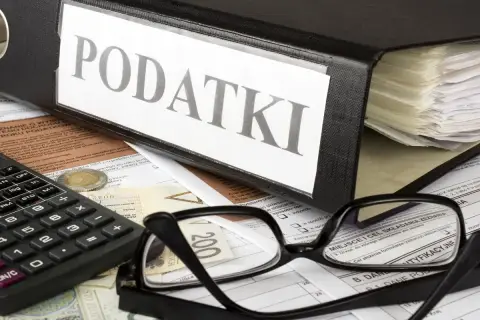 Segregator z napisem "PODATKI", kalkulator, okulary i dokumenty. Obok leży moneta, symbolizująca potencjalną kwotę zmniejszającą podatek.