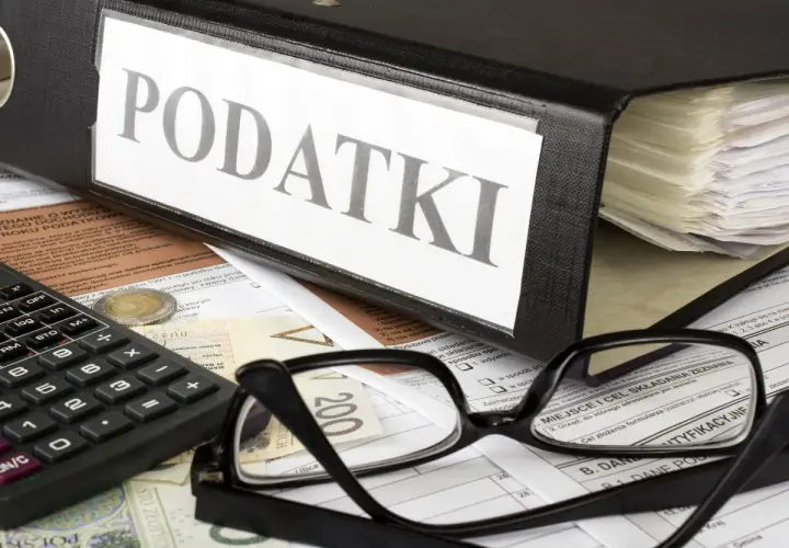Segregator z napisem "PODATKI", kalkulator, okulary i dokumenty. Obok leży moneta, symbolizująca potencjalną kwotę zmniejszającą podatek.