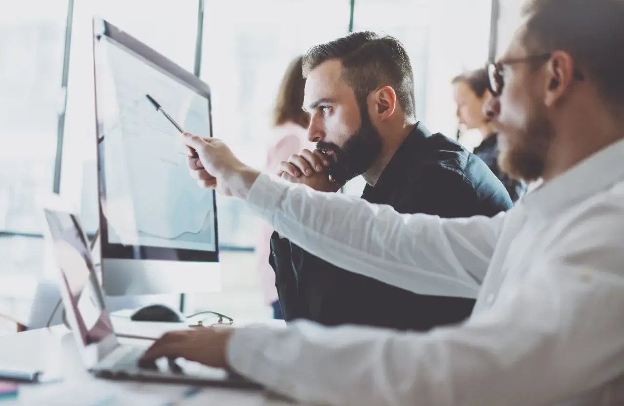 Dwóch mężczyzn analizuje wykresy na ekranie komputera. Jeden z nich, jako business analyst, wskazuje piórem na dane, omawiając je z kolegą.